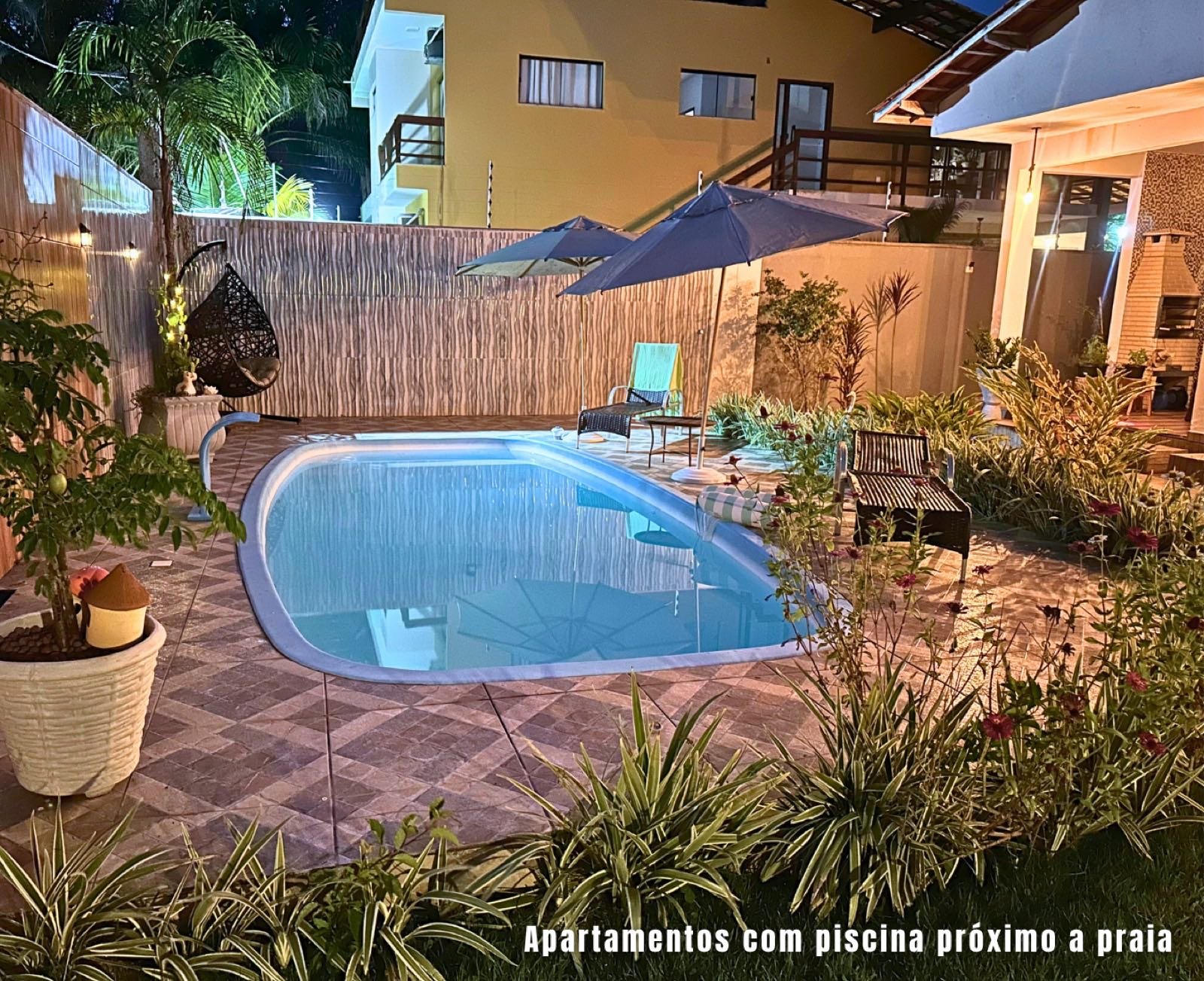 Piscina iluminada à noite com guarda-sóis e jardim na Kitnet Noeli
